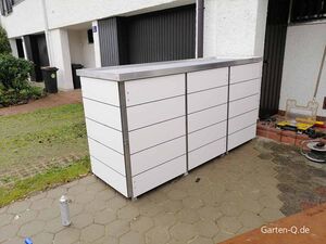 Ein moderner, weißer Gartenschrank mit Metallrahmen steht draußen auf einer gepflasterten Fläche.