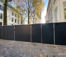 Schwarze Gartenschränke auf einem gepflasterten Gehweg, umgeben von Herbstbäumen und weißen Gebäuden.