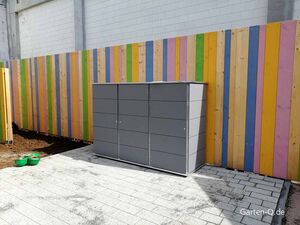 Ein grauer Schrank steht vor einem bunten Holzzaun auf gepflastertem Boden im Garten.