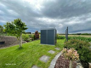 Ein sturmfestes Gartenhaus Garten mit Rasen, Bäumen, Büschen und einem modernen Schuppen unter bewölktem Himmel und starken Wind.