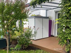 Ein moderner Gartenschrank steht auf einer Holzterrasse, umgeben von Pflanzen und einer überdachten Pergola.