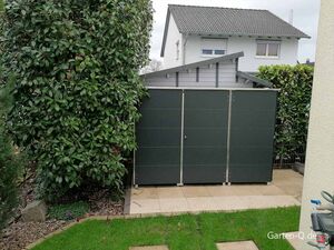 Schuppen im Garten neben grünen Büschen, mit Haus im Hintergrund. Gartenzubehör sichtbar.
